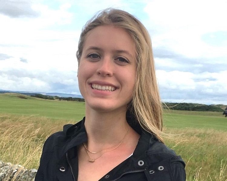 Young woman with long blonde hair, standing in a field of grass. She is wearing a black blouse with an open collar.