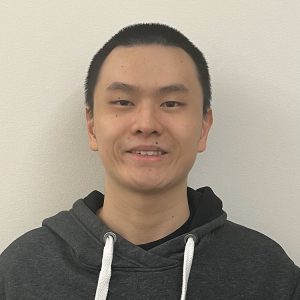 Man with short cropped black hair in front of a white wall, smiling at camera. He is wearing a dark gray hoodie