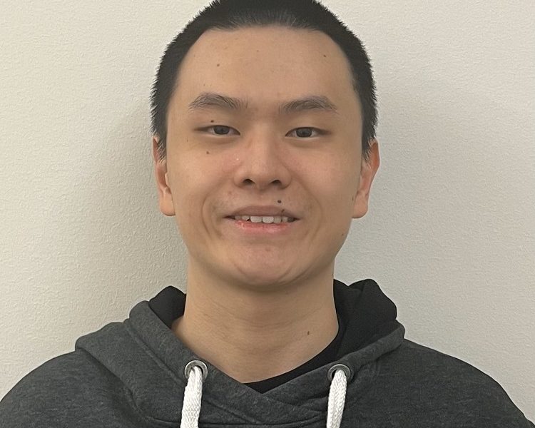 Man with short cropped black hair in front of a white wall, smiling at camera. He is wearing a dark gray hoodie