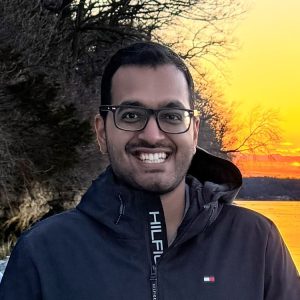 Man standing outside in front of a sunset. He has dark hair and is wearing glasses and a winter coat