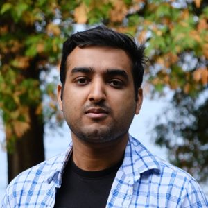 An early career man with dark hair wearing a checkered blue shirt and standing outside.