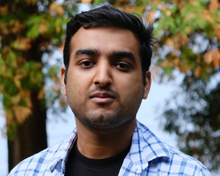 An early career man with dark hair wearing a checkered blue shirt and standing outside.