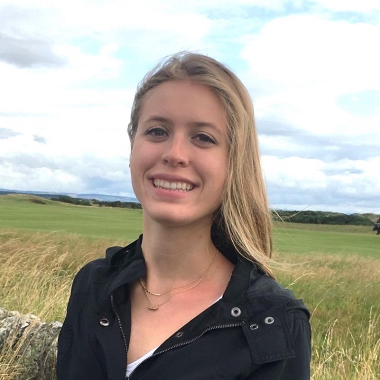 Young woman with long blonde hair, standing in a field of grass. She is wearing a black blouse with an open collar.