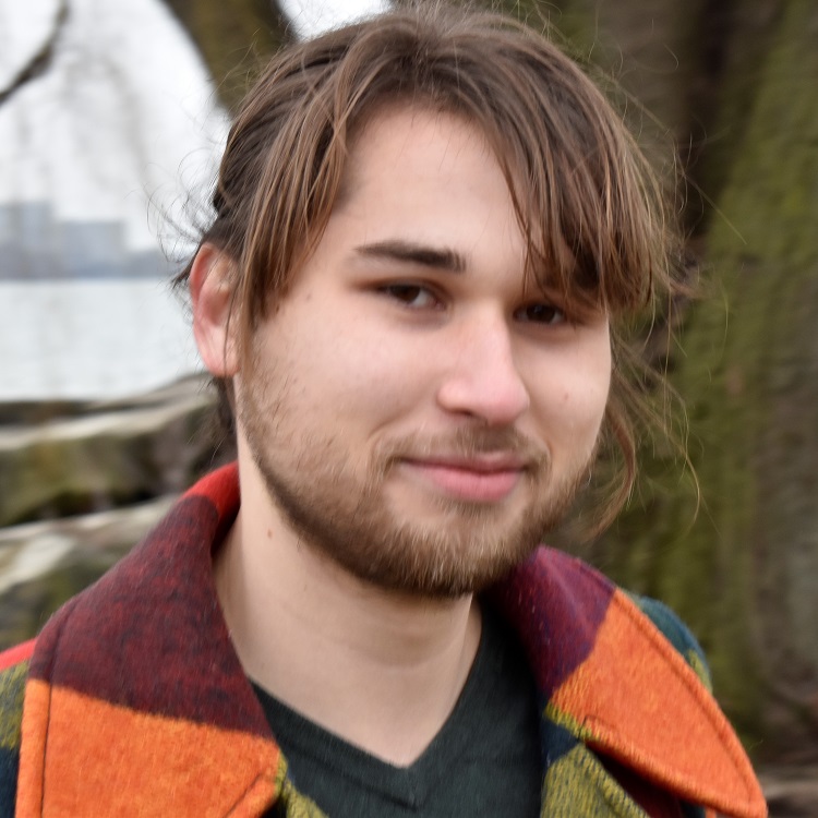 A man with medium-length brown hair wearing a multicolored striped jacket, photographed outdoors.