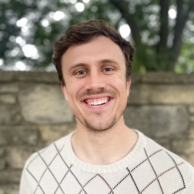 A man with short brown hair and a beard wearing a white patterned sweater, photographed outdoors in front of a stone wall.