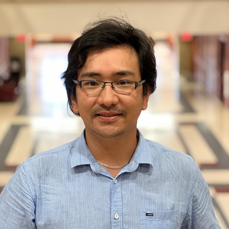 A man with short dark hair wearing glasses and a light blue button-up shirt, photographed indoors in a hallway.
