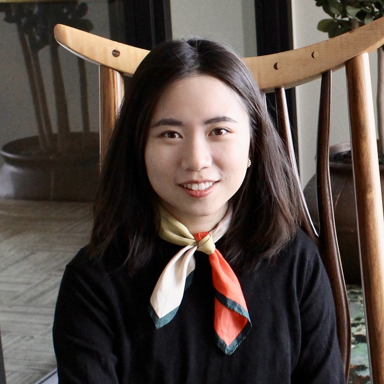 A woman with long dark hair wearing a black top and a colorful silk scarf, seated in front of a wooden chair.
